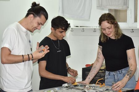 Drei Personen lächeln und arbeiten gemeinsam an einem Bastelprojekt an einem Tisch im Innenbereich.