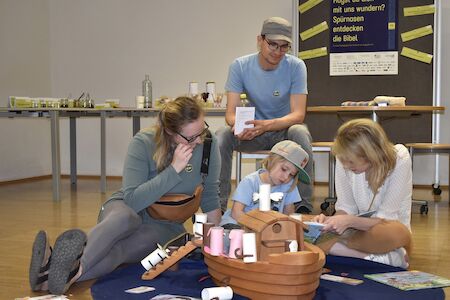 Vier Personen sitzen auf dem Boden und spielen mit einer hölzernen Spielzeugarche und Tieren.