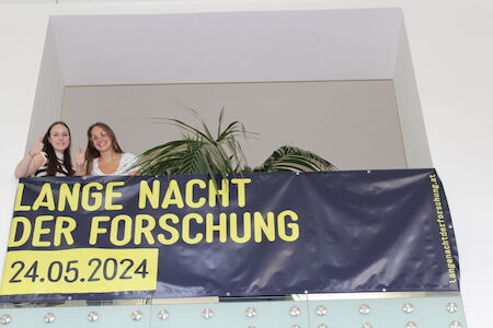 Zwei junge Frauen stehen auf einem Balkon über einem Banner der Lange Nacht der Forschung.