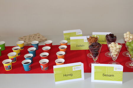 Kleine Tassen mit dunkler Flüssigkeit und beschriftete Schüsseln mit Snacks auf einem Tisch mit rotem Tuch.