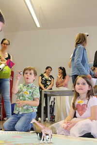 Zwei Kinder spielen auf dem Boden mit Spielzeugtieren, während Erwachsene im Hintergrund stehen.