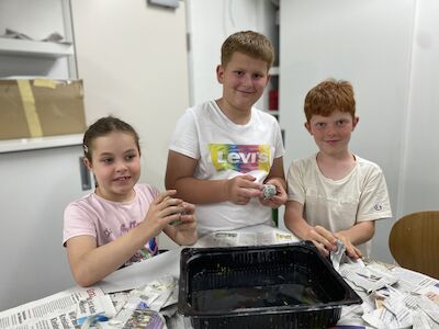 Drei Kinder lächeln, während sie mit Zeitungspapier und einem schwarzen Behälter basteln.