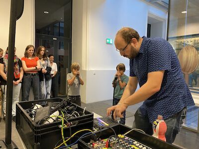 Ein Mann bedient Audiogeräte, während Kinder und Erwachsene in einem Gebäude zuschauen.