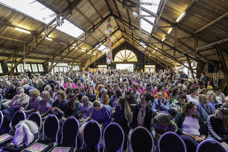 Große Menschenmenge sitzt in einem Holzsaal mit Blick auf die Bühne unter einer hohen Decke.