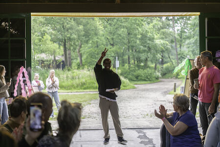 Eine Person steht in einem Türrahmen und zeigt einer Gruppe applaudierender Menschen ein Peace-Zeichen.