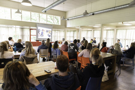 Menschen sitzen in einem Klassenzimmer und sehen sich eine auf eine Leinwand projizierte Präsentation an.