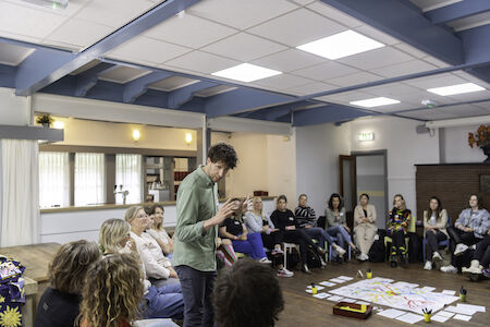 Ein Mann hält vor einer sitzenden Gruppe in einem hellen Raum einen Vortrag, auf dem Boden liegen Unterlagen.