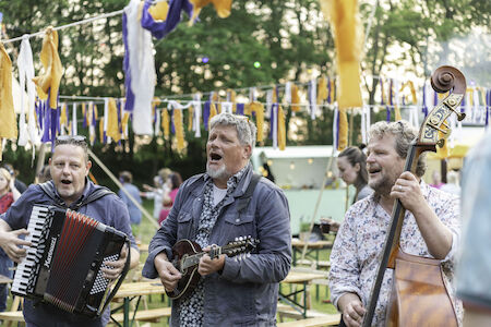 Drei Männer spielen Instrumente und singen bei einem Open-Air-Festival mit bunten Luftschlangen.