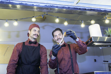 Zwei Männer in roten Hemden und Schürzen bereiten in einer Küche unter Lichterketten Essen zu.