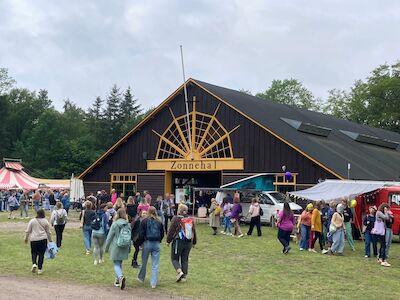 Bei einer Veranstaltung im Freien gehen Menschen auf ein großes Holzgebäude mit der Aufschrift „Zonnehal“ zu.