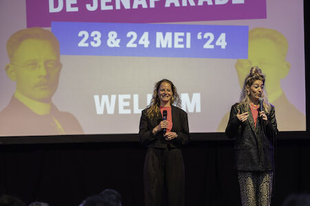 Zwei Frauen sprechen mit Mikrofonen auf der Bühne vor einem Bildschirm mit Veranstaltungsdaten.