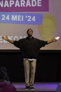 Die Person steht mit ausgebreiteten Armen auf der Bühne, hinter ihr ein Bildschirm mit Veranstaltungsdetails.