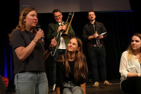 Fünf Personen auf der Bühne; eine Frau spricht in ein Mikrofon, andere halten Musikinstrumente.