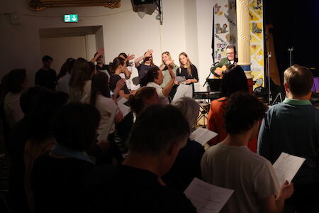Eine Gruppe von Menschen singt gemeinsam in einem Gebäude, einige halten Textblätter in den Händen.