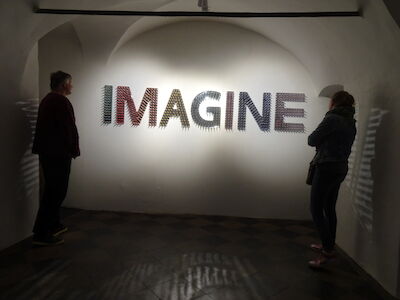 Zwei Personen stehen da und betrachten eine Wandkunstinstallation, auf der „IMAGINE“ steht.