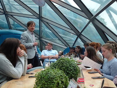 Ein Mann steht in einem Glasraum und spricht zu einer Gruppe, die um einen Tisch herum sitzt.