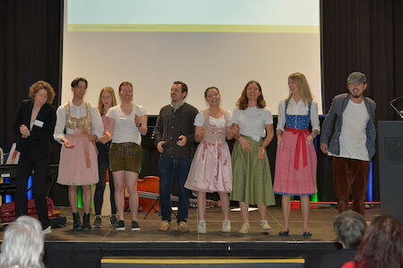 Eine Gruppe von Menschen in traditioneller und legerer Kleidung steht lächelnd auf der Bühne.