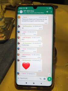 Auf einem Smartphone wird ein WhatsApp-Gruppenchat mit Nachrichten und einem roten Herz-Emoji angezeigt.