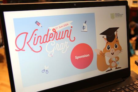 Auf dem Laptop-Bildschirm ist die Kinderuni Graz mit einem Cartoon-Eichhörnchen mit Abschlusskappe zu sehen.