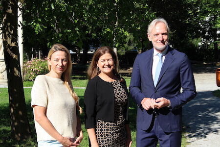 Drei Personen stehen an einem sonnigen Tag draußen und lächeln in die Kamera.