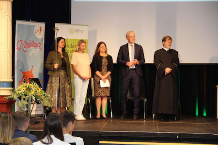 Bei einer Veranstaltung stehen fünf Personen auf der Bühne und blicken auf ein Publikum.