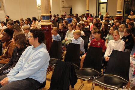 Eine Gruppe Kinder sitzt bei einer Indoor-Veranstaltung aufmerksam in Stuhlreihen.
