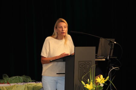 Frau spricht an einem Podium mit Mikrofon, gelbe Blumen im Vordergrund.