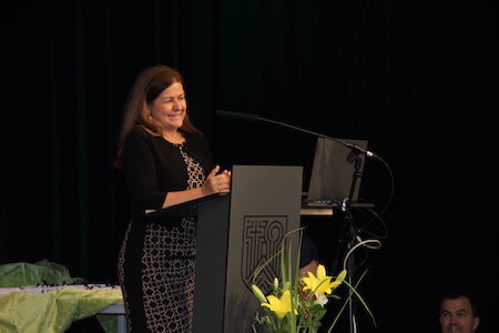 Eine Frau steht lächelnd auf einem Podium, im Vordergrund sind gelbe Blumen zu sehen.