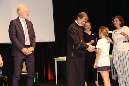 Ein Mann im Bademantel überreicht einem jungen Mädchen auf der Bühne eine gerollte Urkunde.