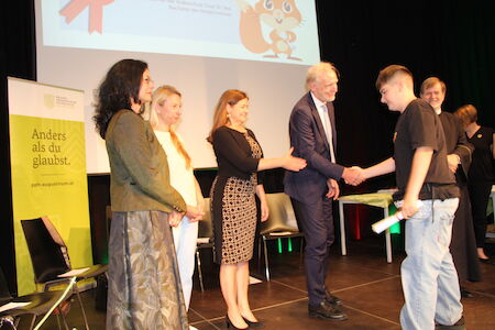 Ein Mann schüttelt einem jungen Preisträger auf der Bühne die Hand, während andere zuschauen.