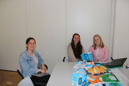Drei Frauen sitzen mit Laptop, Snacks und bunten Papieren an einem Tisch.