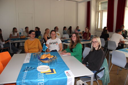 Vier Frauen sitzen an einem Tisch mit Gebäck, weitere sitzen an Tischen im Hintergrund.