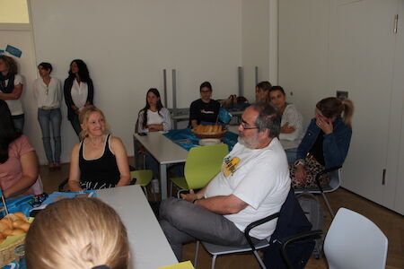 Menschen sitzen in einem Raum um Tische herum, manche unterhalten sich, andere hören zu.