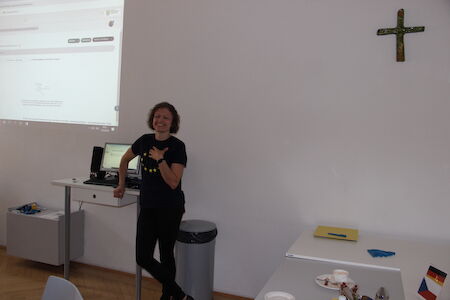 Frau steht an einem Schreibtisch in einem Raum mit einer Projektionsfläche und einem Wandkreuz.