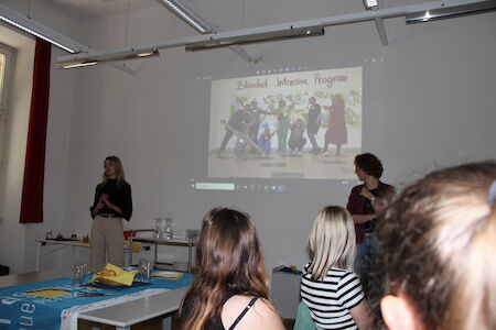 In einem Klassenzimmer mit mehreren sitzenden Teilnehmern sehen sich Personen eine projizierte Präsentation an.