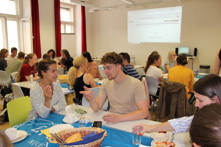 Menschen sitzen an Tischen, unterhalten sich und essen in einem hellen Klassenzimmer.