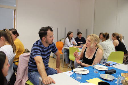 Menschen sitzen an Tischen, unterhalten sich und essen in einer entspannten Innenatmosphäre.