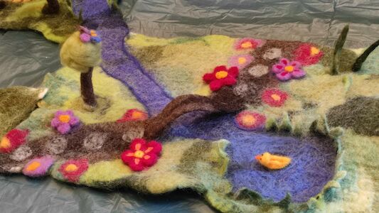 Filzlandschaft mit einem Fluss, Blumen, Bäumen und einem kleinen gelben Vogel.