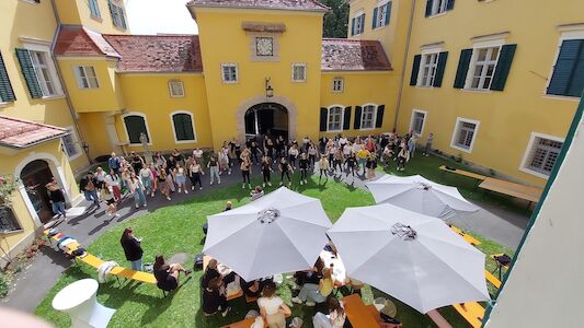 Eine Menschenmenge versammelt sich in einem sonnigen Innenhof mit gelben Gebäuden und weißen Sonnenschirmen.