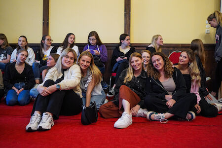 Eine Gruppe junger Frauen sitzt lächelnd zusammen auf einem roten Teppich in einem Raum.