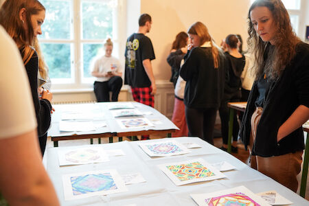 Menschen betrachten farbenfrohe Kunstwerke, die auf Tischen in einem hell erleuchteten Raum ausgestellt sind.