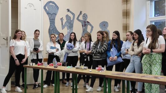 Eine Gruppe junger Frauen steht in einem Klassenzimmer um Tische mit verschiedenen Gegenständen herum.
