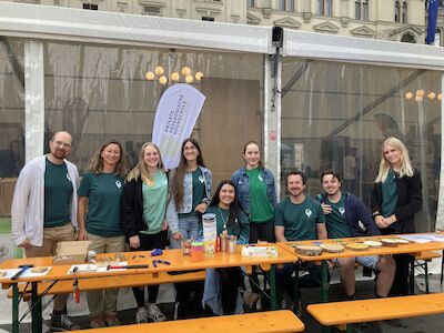 Neun Personen in grünen Shirts posieren im Freien hinter einem Tisch mit Bastelmaterialien.