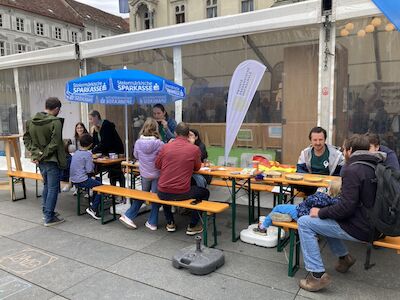 Menschen sitzen an Tischen im Freien unter einem Zelt, unterhalten sich und essen zusammen.