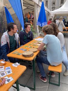 Vier Personen sitzen mit Karten und Holzklötzen an einem Tisch im Freien und unterhalten sich.