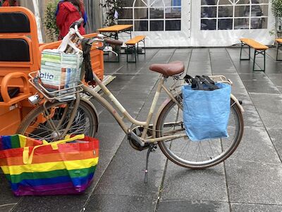 Fahrrad mit Einkaufstüten, darunter eine Regenbogentasche, auf nassem Asphalt geparkt.