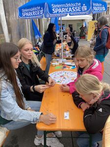 Unter einem Sparkassenzelt sitzen vier Mädchen an einem Tisch im Freien mit Bastelutensilien.