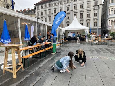Zwei Mädchen spielen auf dem Bürgersteig neben Tischen und Zelten auf einem Stadtplatz.