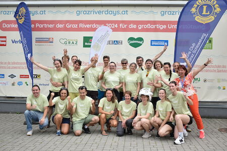 Eine Gruppe von Menschen in passenden Hemden posiert und lächelt im Freien, hinter ihnen sind Banner zu sehen.