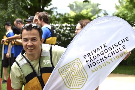 Lächelnder Mann in einer Schwimmweste neben einem Banner der Privaten Pädagogischen Hochschule Augustinum.